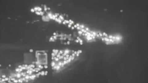 Grainy/black and white blurry image of lots of car lights on a congested M11 going under a bridge 