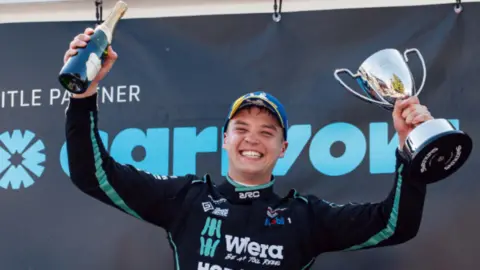 A man mainly wearing black is smiling, standing up and holding a trophy in his left hand and a bottle in his right hand. The mainly black background includes company names.
