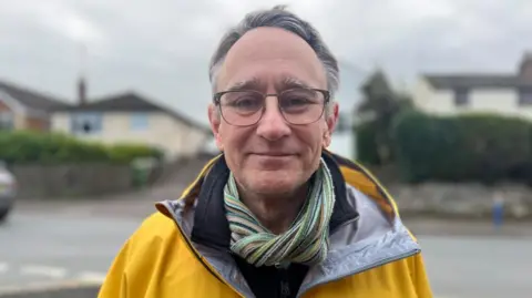 Leckhampton councillor Martin Horwood. He has grey hair and glasses smiles at the camera while wearing a green and brown stripy scarf, and a yellow raincoat with grey lining. He's standing in front of a Church Road with housing and hedges blurred in the background. 