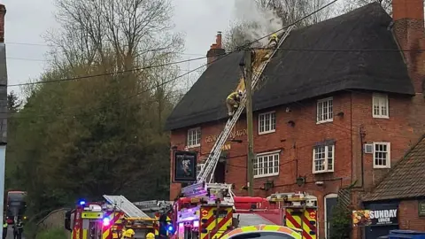 A red brick Inn is seen with two fire engines parked outside. Firefighters are climbing up a ladder on to the roof.