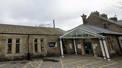 Google The entrance to Oxenholme train station. It is a one-storey stone building. There is a sign reading Oxenholme Lake District next to the entrance. 