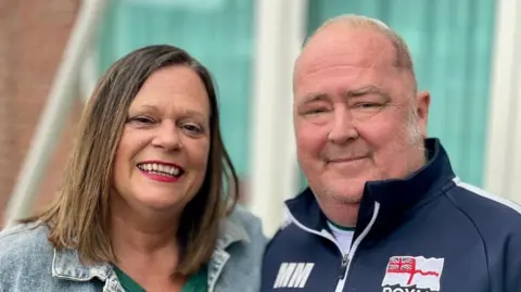 Steve Huntley/BBC Debbie Monaghan is on the left wearing a green top and a light blue denim jacket. She has shoulder length brown hair and is looking towards the camera smiling. On the right is Mark Monaghan who is wearing a navy jacket with a white top, and is looking towards the camera smiling.