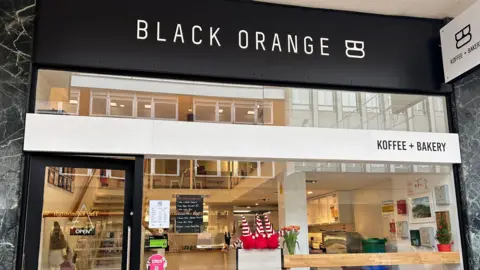 The outside of the Black Orange, which has a black sign with white capitals. Underneath is a white sign reading Koffee + Bakery. It has a glass frontage behind which a counter can be seen and stairs to a second level with further seating.