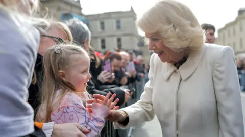 Queen Camilla is meeting a little girl with her mother behind a low metal fence. The queen is smiling and hold the girls hand. The girl is smiling back at her wearing a pink jumper and two pony tails. 