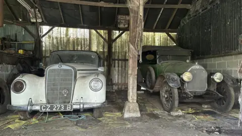 Two dusty cars - a white Mercedes from the 1950s and a 1921 green Talbot - pictured with bonnets facing the camera in a dilapidated barn