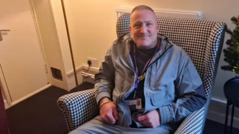 Jeremy Palmer sitting in a wing-backed black-and-white chequered chair in a plain room with beige walls and a brown carpet. He is smiling and wearing a lanyard and a grey tracksuit.