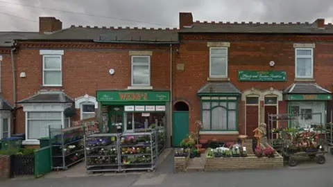 Google The front of a shop, which has green signage above the door, has racks of flowers outside. It is situated among a row of terraced houses.