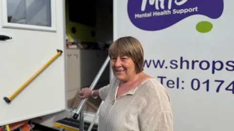 A woman, who has short brown hair and is wearing a beige top, is holding onto a metal rail that leads up some steps into a white bus.
