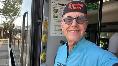 A man wearing a cycling jersey stands in the open doorway of a train at a station platform, with the train visible beside them.
