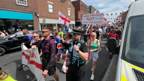 A demonstration waving England flags walking down a high street lined by brick buildings. One placard is visible, which reads "Illegal immigrants are not welcome. Stop the boats."