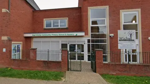 Google A red brick school with large long windows and dark railings and patches of grass on either side of the gates. A sign above the reception says "East Wichel Community Primary School and Nursery".  A sign to the left also has writing on. 
