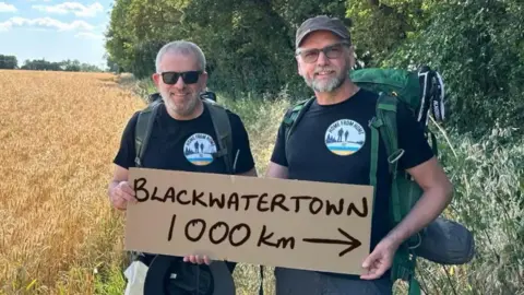 Standing at the edge of a field of golden grain, two men hold a cardboard sign that reads: 'BLACKWATERTOWN 1000km'. An arrow on the sign points in the direction of trees that line the field.