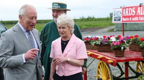 King Charles III, wearing a grey suit, standing beside an older man and woman outdoors. There is a cart with a few flower pots on top of it. 