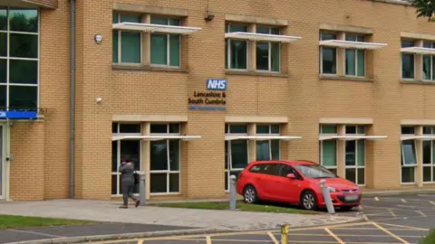 Google The headquarters of Lancashire and South Cumbria NHS Foundation Trust (LSCFT) in Preston. It is a two-storey beige brick building with windows . A person is walking towards the reception. A red car is parked in front of the building. 