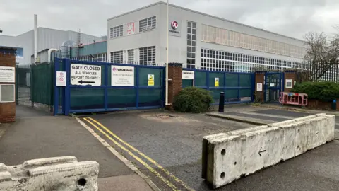 Two-storey factory building, mainly grey with a smaller blue section between two larger buildings. The entrance is blocked by concrete barriers. There are blue gates beyond the barriers, with direction notices attached.