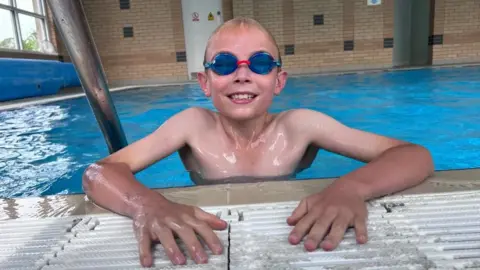 Nine year old Mateo is in a swimming pool at the edge, looking into the camera lens and smiling. He is wearing blue goggles.