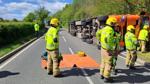 An orange lorry is on its side at the side of a main road through countryside. Several firefighters in uniform and yellow helmets are around it.