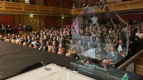 Jon Wright/BBC An auditorium full of people with a glass trophy in the foreground.