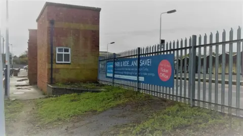 Kettering Civic Society An area of a railway station without plants, showing metal fencing, brick structures and a ramp with glass and mud on it. A flower bed, in the corner is empty. There is a sign on the railings. 