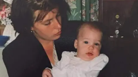 A woman with short dark hair holds a baby in a white dress.