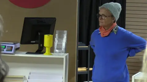 Jo on the stage, where a computer, collection tin and scales are visible on a shelf.