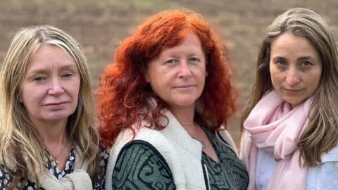 Three women standing in front of brown land in a field. On the left the woman has dark blonde hair and a black and white top. In the middle, the woman has curly red hair and a green and black patterned top with a white gilet. On the right the woman has dark blonde hair with a white jumper and pink scarf on 