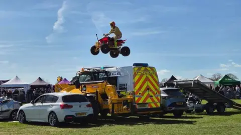 A man on a quad bike in mid-flight while jumping over two cars, a digger and a police van. It is a sunny day at a show and there are marquees in the background. The stuntman is wearing a yellow suit, red and yellow helmet and his quad bike is yellow and red. A ramp is propped up before the first car.