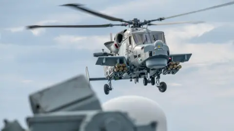 A grey Royal Navy Wildcat helicopter in flight, equipped with the Martlet missile system. The picture is a close up and the helicopter's rotor's are spinning. 