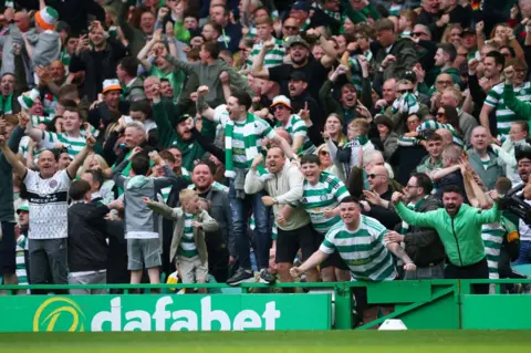 Reuters A packed crowd of Celtic supporters celebrate wildly in the stands during a Scottish Premiership match at Celtic Park. Fans wearing green-and-white striped shirts and scarves cheer, raise their fists, and embrace as excitement spreads through the stadium.