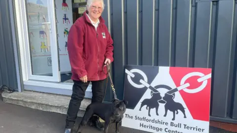 A woman in a red fleece jacket and black trousers stands looking towards the camera and smiling. She has a black Staffordshire bull terrier on a leash which is standing at her feet. They are standing in front of the doorway to a building which is clad in corrugated metal. Next to them is a red, black and what sign that says The Staffordshire Bull Terrier Heritage Centre.