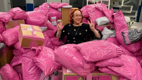 Rachel Grady sits among a large pile of pink parcels. She has a ginger curled bob and holds two peace signs up to either side of her head. She smiles widely and wears pink glasses.