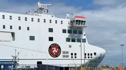 The bow of the Manxman ferry moored in Douglas Harbour. It is a white vessel with the Isle of Man Steam Packet Company's logo on the side, which features a white three-legs-of-Man symbol in a red circle with a black band around it featuring the company's name in white lettering.