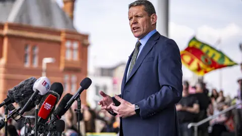 Rhun ap Iorwerth in a dark blue suit and a light blue shirt, wearing a dark green and pink dotted tie standing on the steps of the Senedd