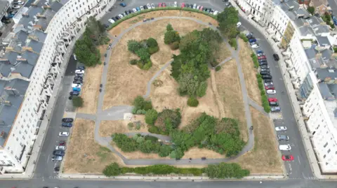 Eddie Mitchell An aerial view of a city square. There is a horseshoe shape of white, Regency-style housing surrounding a park. There are a few green trees in the park but the grass is parched and brown.