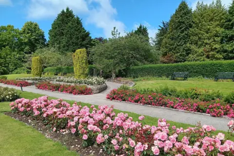 Colin Dey A manicured park with planted pink and red flowers. A gravel path runs around the neat flowerbeds and benches sit on the grass in the distance in front of a hedge.