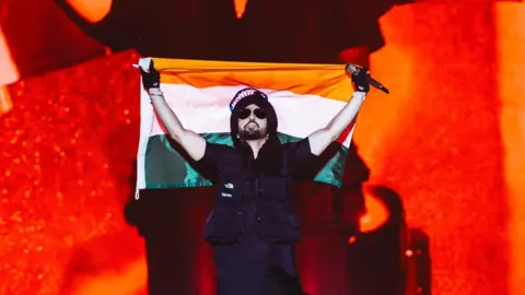 A picture of artist Diljit Dosanjh wearing a black Indian traditional shirt with a black jacket, sunglasses and a black turban holding the Indian flag and a mic during his concert in Delhi.