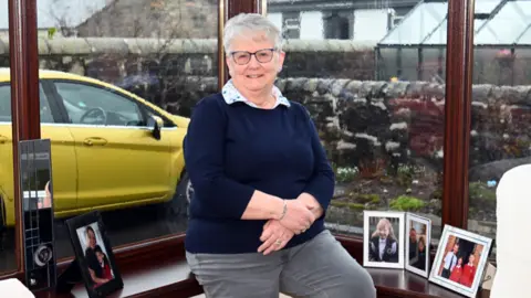 Charlie Gilmour/BBC A woman perches on the windowsill of her bay window, wearing a navy jumper and grey trousers. There are framed photographs of her children and grandchildren around her and a yellow car is visible through the window.