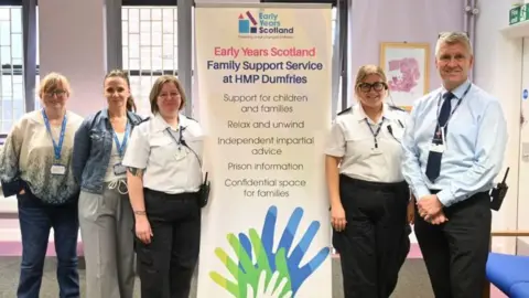 Scottish Prison Service Four adults stand around a sign describing the Early Years Scotland Family Support Service at HMP Dumfries.