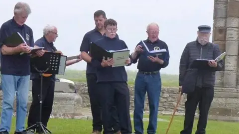 Men of Staithes Six men in navy and black, from the Men of Staithes fishermen's choir, perform at a festival in Whitby Abbey. They are standing on grass with the stone pillars of Whitby Abbey behind.