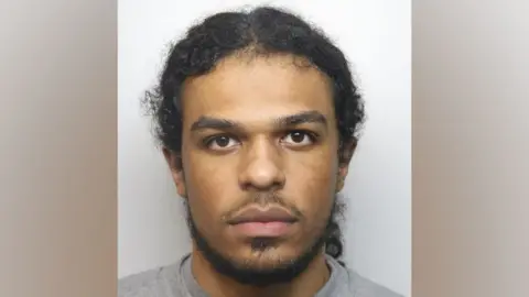 Thames Valley Police Jayden Gubetini mugshot pictured looking straight at the camera, not smiling. He has a goatee beard and hair tied back in a ponytail, brown eyes and wearing a grey sweater.