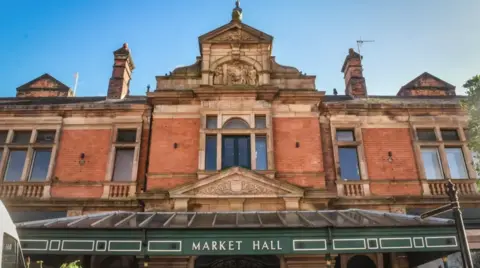 East Staffordshire Borough Council The entrance to Burton Market Hall, with a glass canopy fixed to a red brick building with ornate architectural features. There is white lettering on the canopy which reads 'Market Hall' and there are windows on a second floor of the building.