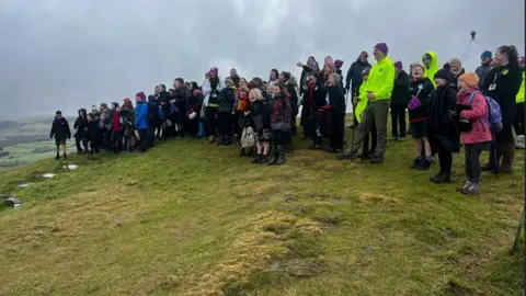 A crowd of children and adults wearing walking gear on a hill 