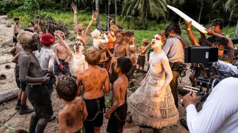 BBC/Eleven/J Redza A group of boys during the filming of Lord of the Flies on an island. They have body paint on them and mud. They are dancing and shouting as they are filmed. 
