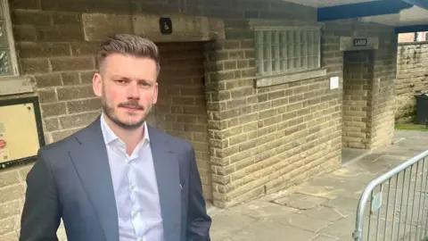LDRS A man with short dark hair wearing a white shirt and grey suit jacket stood outside a public toilet