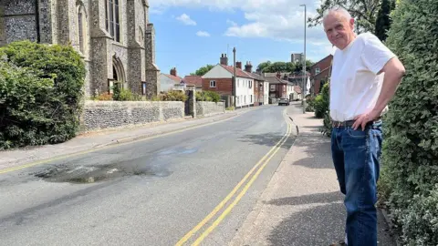 Ian Clark stands on the pavement on Cromer Road looking at a wet, bumpy patch of road which has been damaged by a water leak. Ian is wearing a white t-shirt and blue jeans. He has his glasses on his head. 