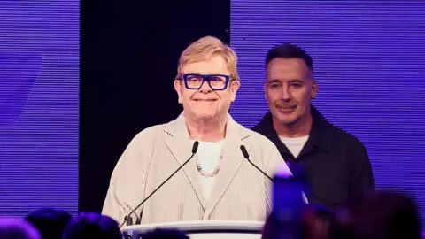 Getty Images Elton John appears at a podium in a light coloured blazer, white top and blue glasses. His husband David Furnish stands behind him. They stand in front of a digital screen. 
