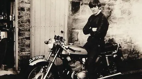 Gordon Castle A black and white image of Gordon Castle in his 20s on a 1970s motorbike wearing a black top parked in front of a wooden door. His arms are folded and he is unsmiling.
