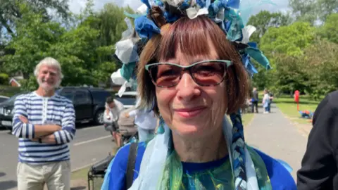 Dee is stood by the river with other campaigners and smiling at the camera. She is wearing a blue and green top and has a blue and white decorated hairband on her head.