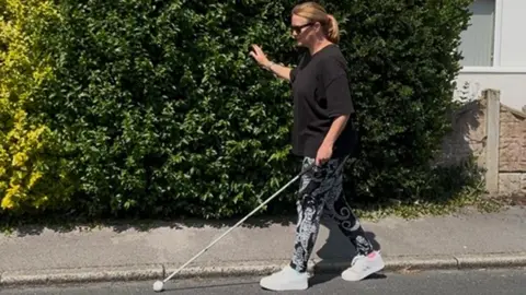 Lorraine walking down a street by a large hedge in dark glasses and with a white cane