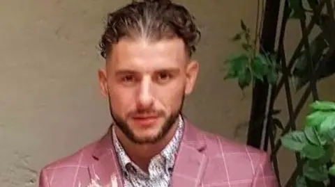 This photo shows a person in what appears to be an outdoor courtyard or garden setting.
The seated person occupying most of the center of the photograph.
The person is wearing a distinctive dusty pink suit with a subtle white windowpane check pattern.
Under the suit jacket is a light-colored patterned dress shirt.
The person has short, styled hair and a neatly trimmed beard.